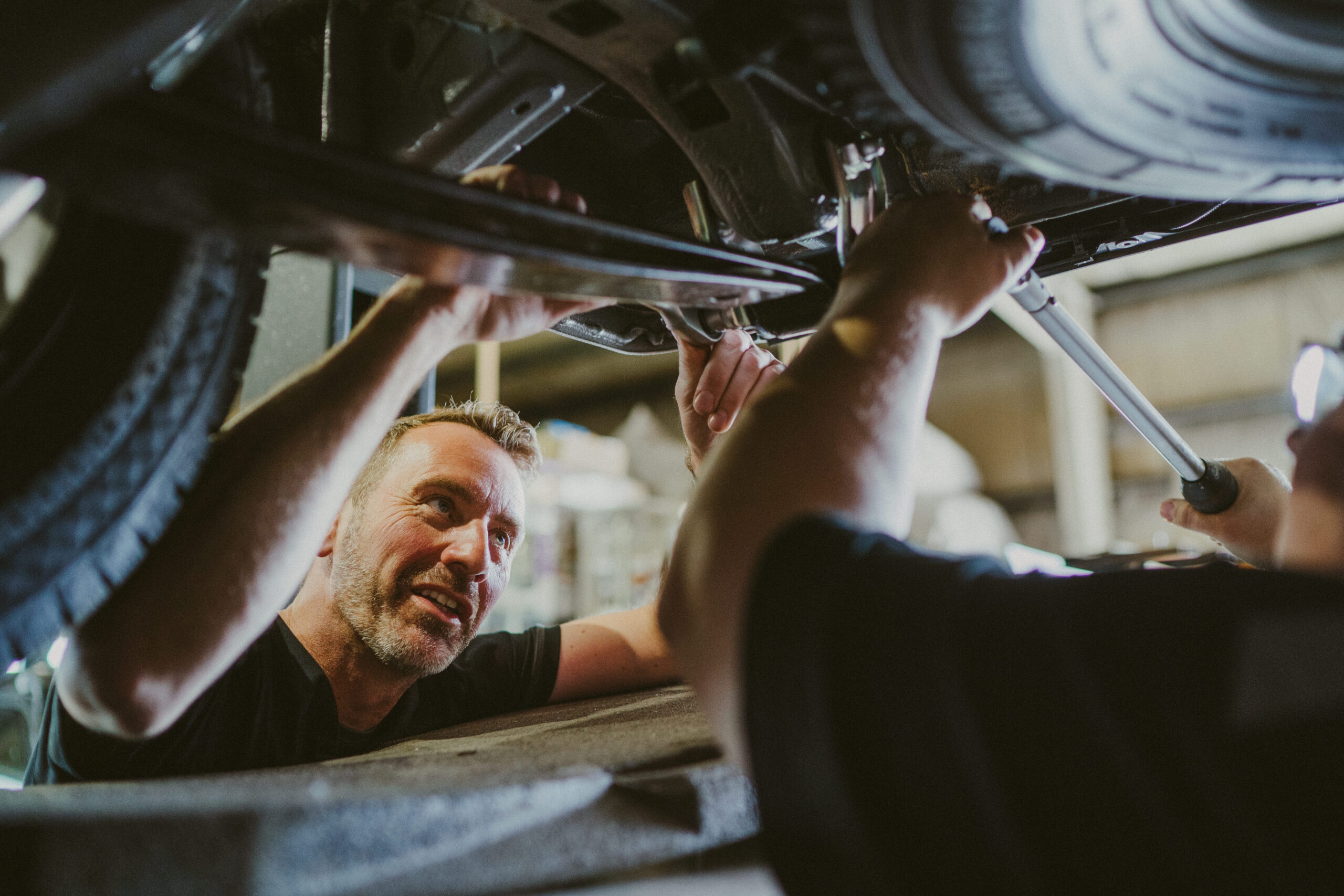 Mechaniker arbeitet unter einem Fahrzeug und blickt konzentriert zu einem Kollegen, während beide gemeinsam an einer Reparatur arbeiten.