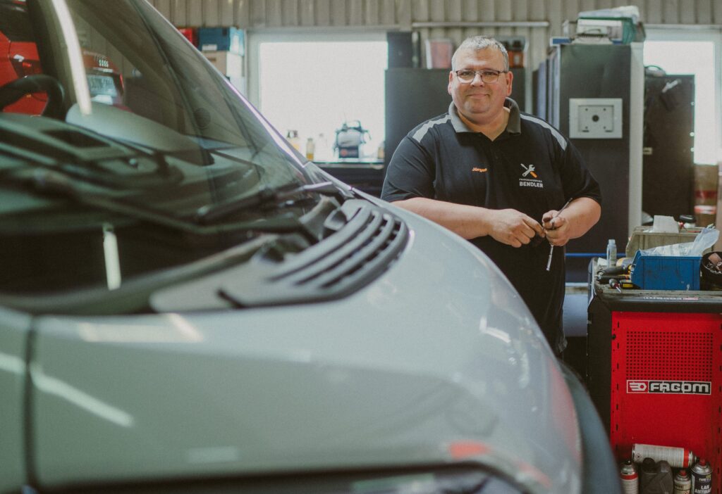 Mechaniker in der Werkstatt von Fahrzeugservice Bendler, der an einem Campervan arbeitet und ein Werkzeug in der Hand hält.